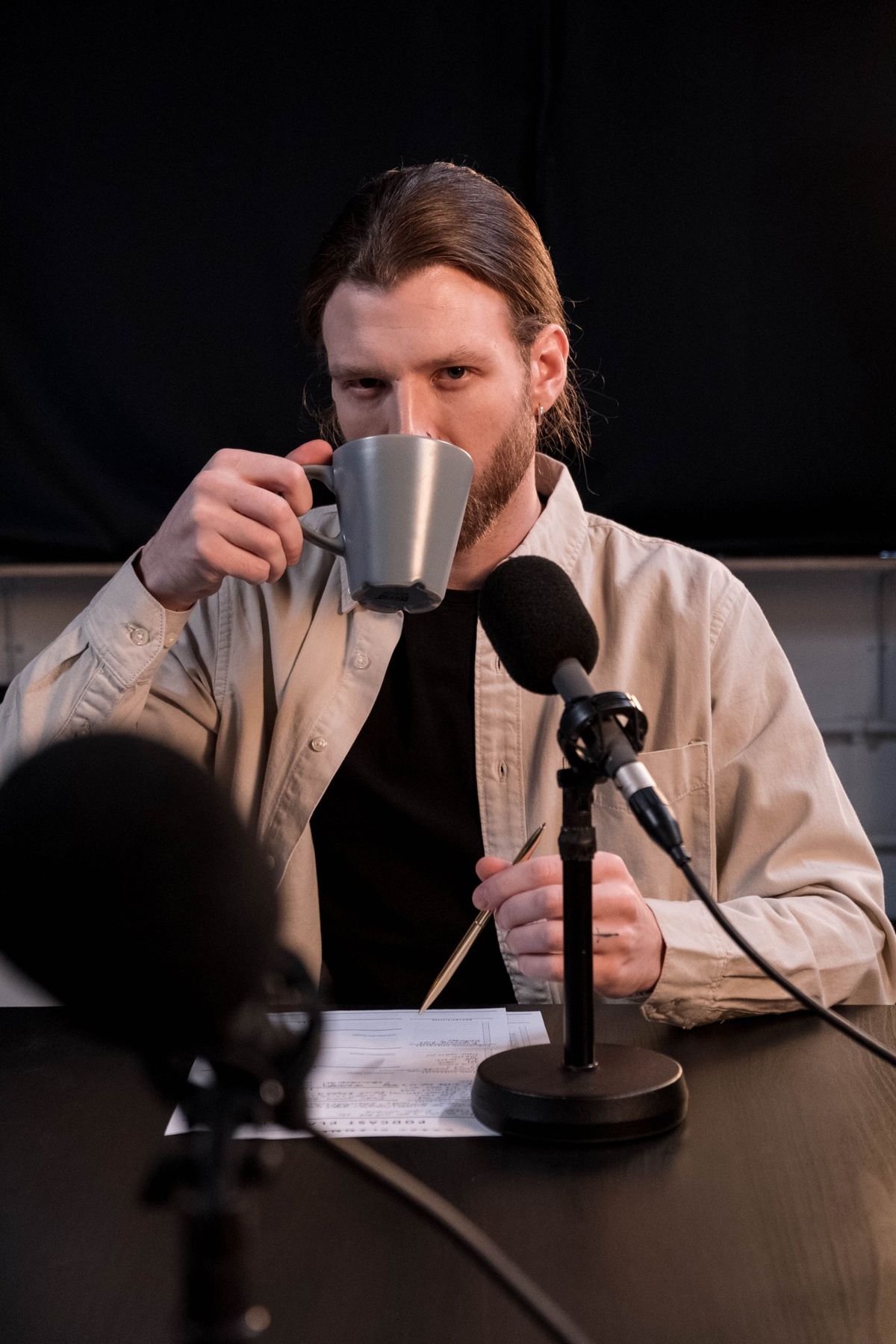 A presenter sipping coffee between two microphones in a dark studio