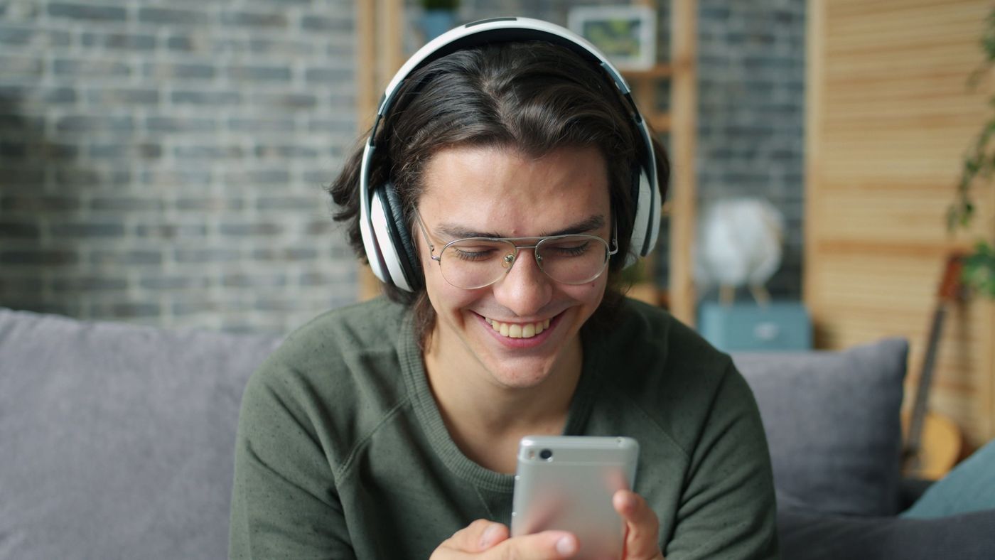 A smiling listener in headphones scrolling his phone on a sofa
