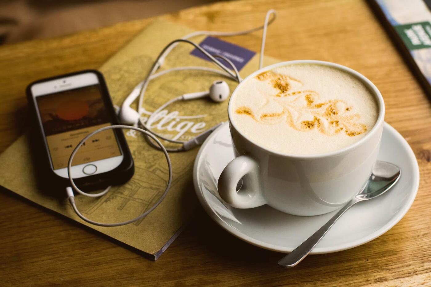 A phone playing a station next to a cappuccino and earphones