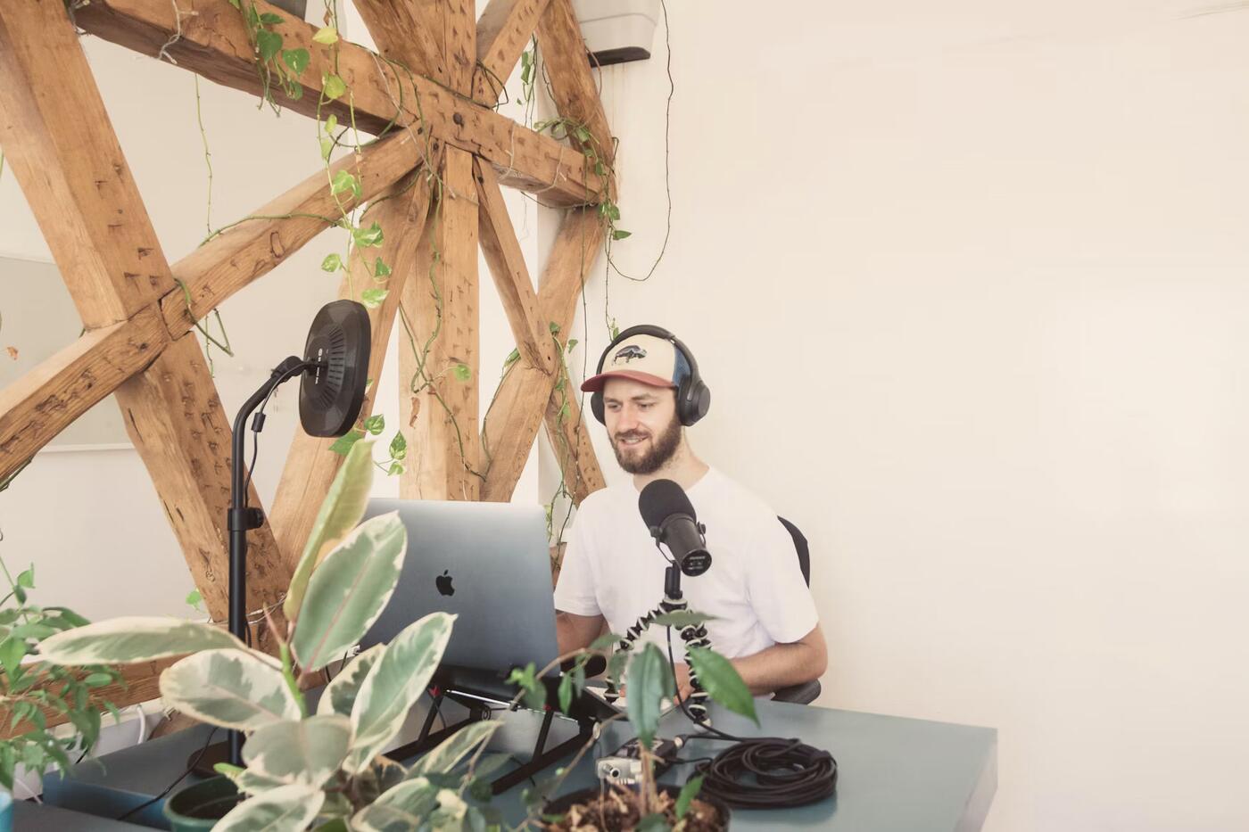 A broadcaster with a microphone and laptop in a bright studio full of plants