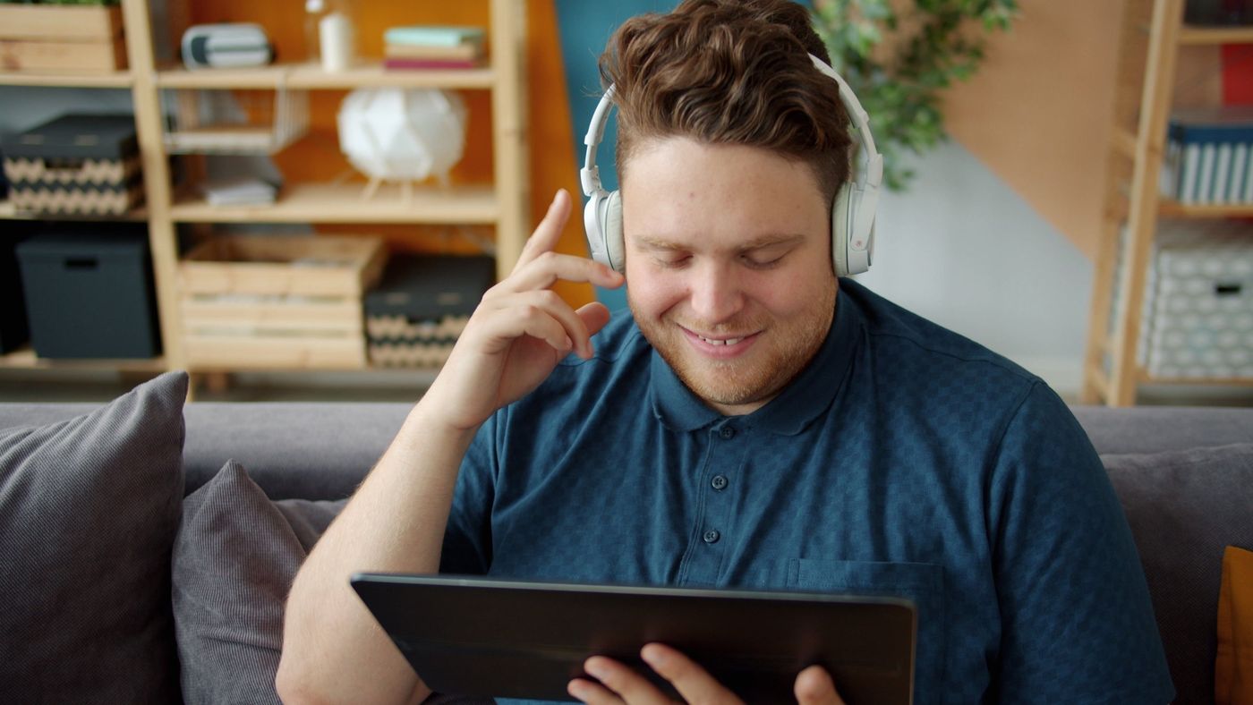 A man in white headphones smiling at a tablet on his sofa